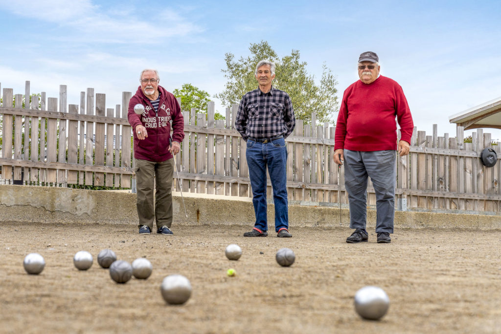 Trois hommes jouent à la pétanque sur terrain sableux.